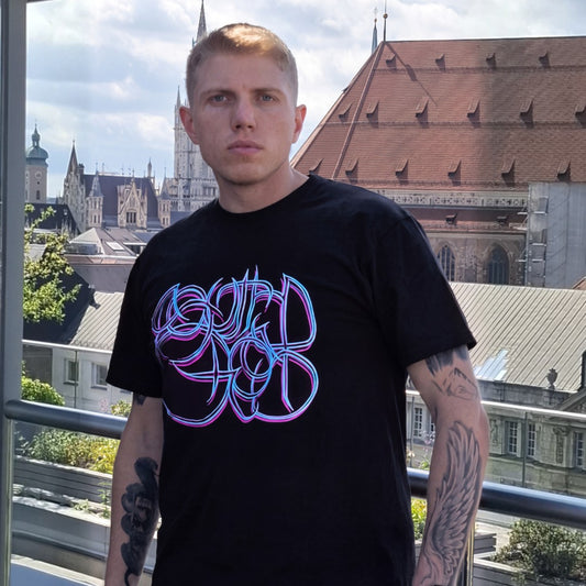 Sascha Stammer Wurth de Rothschild with tattooson hos arms, stands on a balcony wearing a black t-shirt with a purple blue design by BOB B.  on the chest, with a cityscape of Munich in the background background.