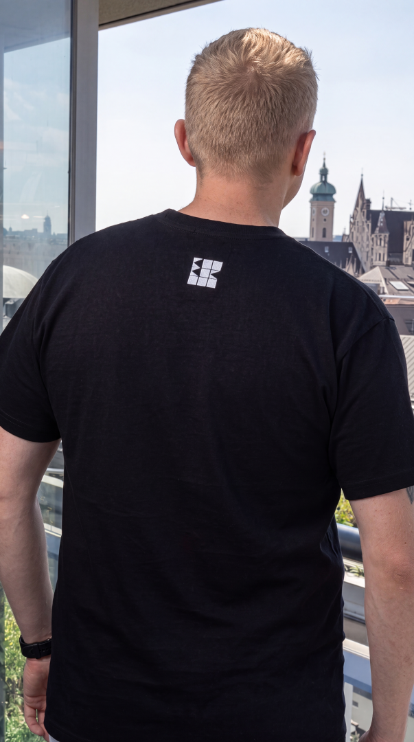 Sascha Stammer Wurth de Rothschild with tattoos on his arms, stands on the balcony of Bayrischer Hof wearing a black BBB Collection t-shirt with a purple blue South Ghost design by BOB B.  on the chest. Cityscape of Munich in the background.