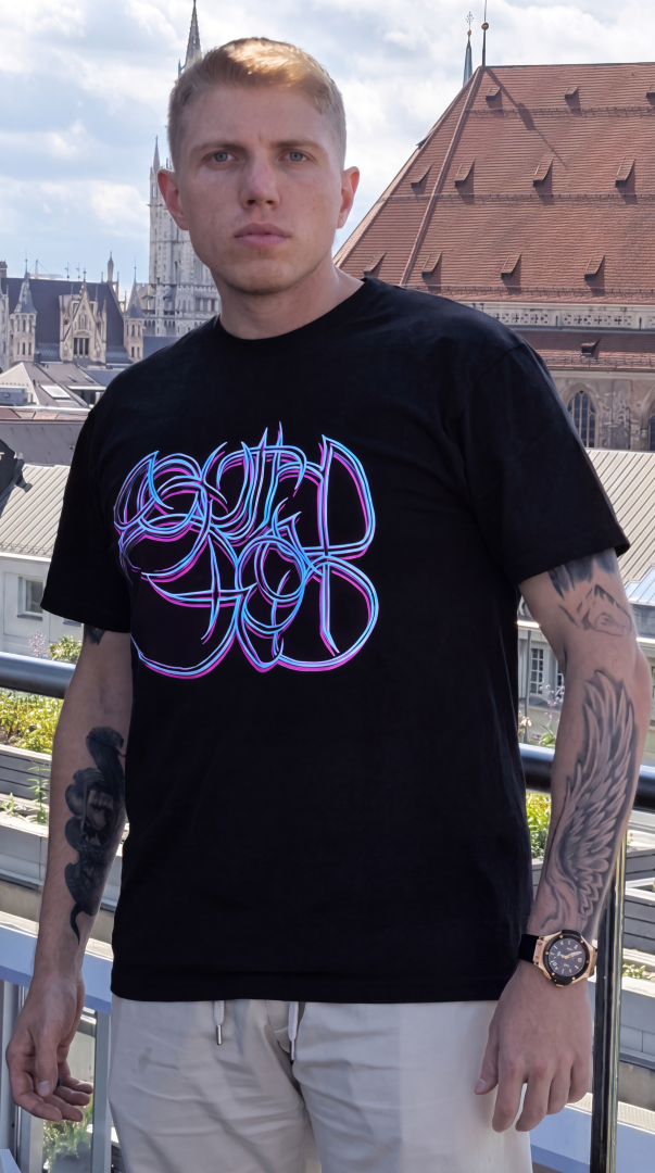 Sascha Stammer Wurth de Rothschild with tattoos on his arms, stands on the balcony of Bayrischer Hof wearing a black BBB Collection t-shirt with a purple blue South Ghost design by BOB B.  on the chest. Cityscape of Munich in the background.