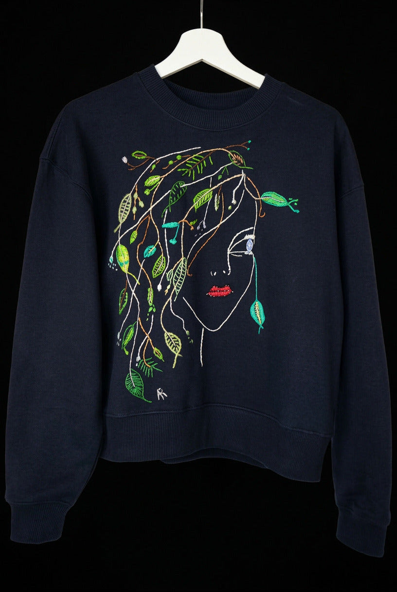 A dark navy blue crewneck sweatshirt featuring a stylized graphic of a woman's face with green leaves and vines woven through her hair, hanging on a white hanger 