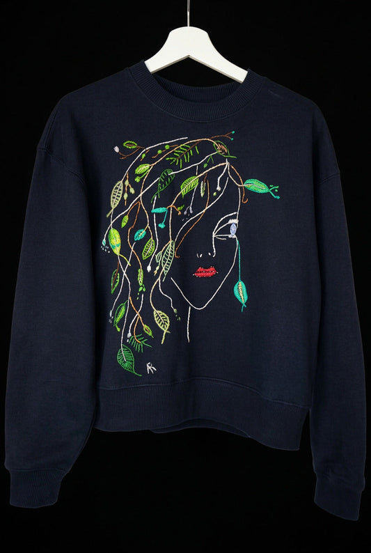 A dark navy blue crewneck sweatshirt featuring a stylized graphic of a woman's face with green leaves and vines woven through her hair, hanging on a white hanger 