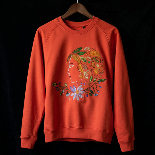 An orange crewneck sweatshirt featuring a large, colorful embroidered graphic of a woman's profile with floral and leaf details on the front, displayed against a textured gray wall.